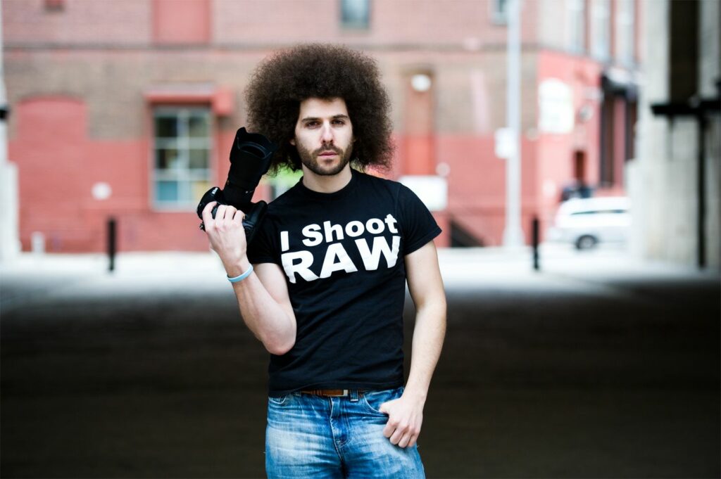 An image of pro photographer Jared Polin holding a camera, and wearing one of his signature, "I Shoot RAW" t-shirts.
