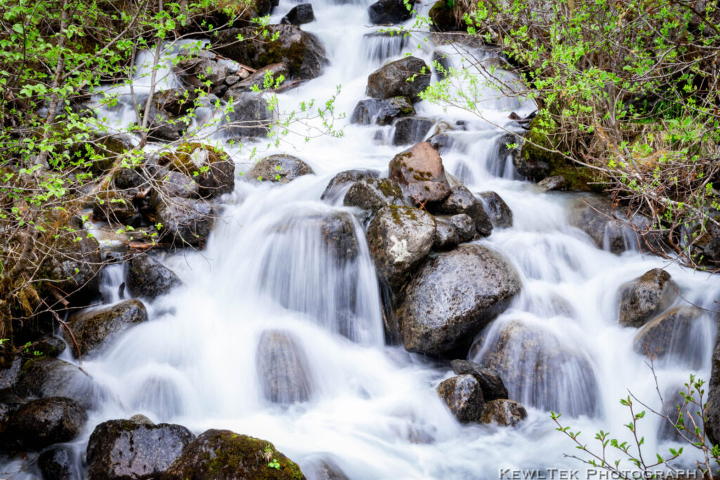 Image of water flowing over large boulders in a woodland stream with a silky looking waterfall effect.