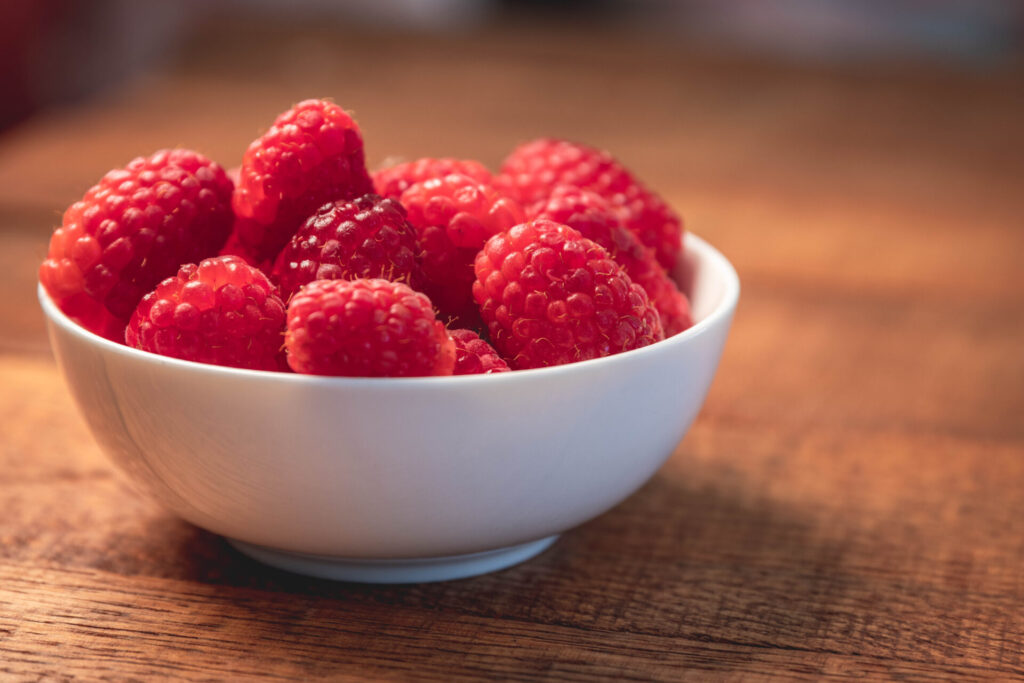 Photo of a white bowl filled with juicy red raspberries, and lots of smooth background blur.