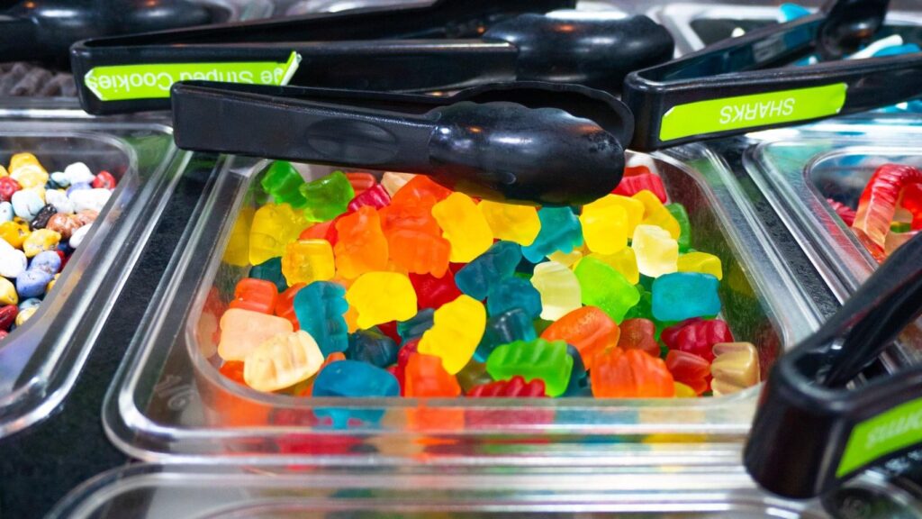 Photo of a container of Gummy Bears on a frozen yogurt toppings bar, with tongs, and other toppings in the frame. 