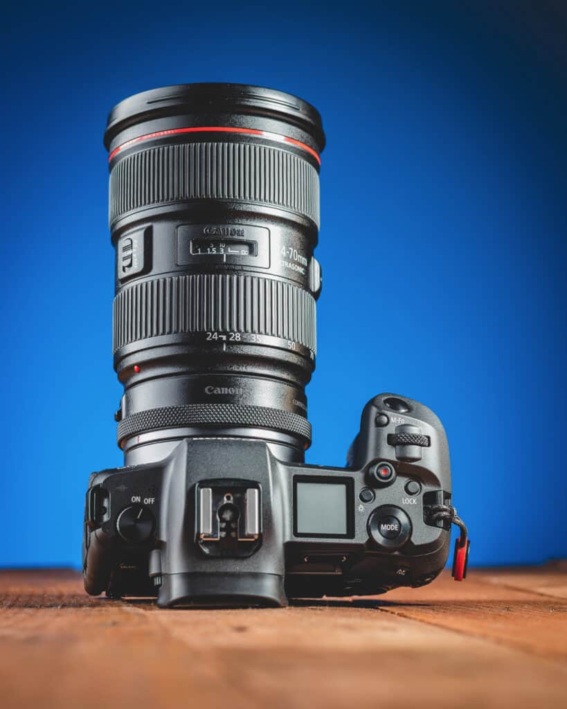 Canon EOS R camera with Canon EF 24–70mm f/2.8 L zoom lens attached using an RF-to-EF adapter, product shot on wooden table with blue background.