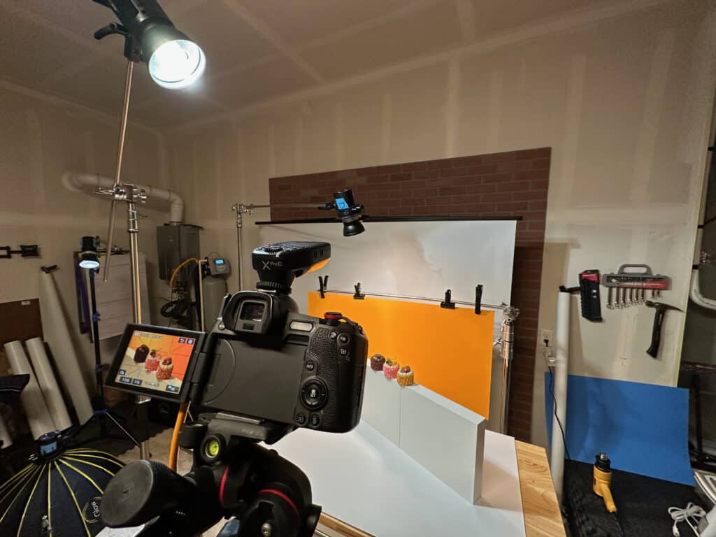 Behind-the-scenes view of a home studio product photography setup using portable Godox strobes, camera on tripod, and colored background panels.