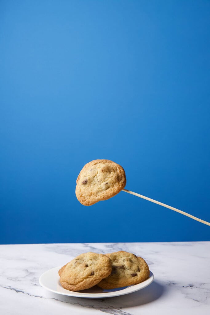 Single chocolate chip cookie photographed midair on a wooden skewer, positioned above a plate to be composited into a levitation image.
