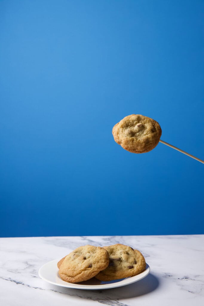 Single chocolate chip cookie photographed midair on a wooden skewer, positioned higher above a plate to be composited into a levitation image.
