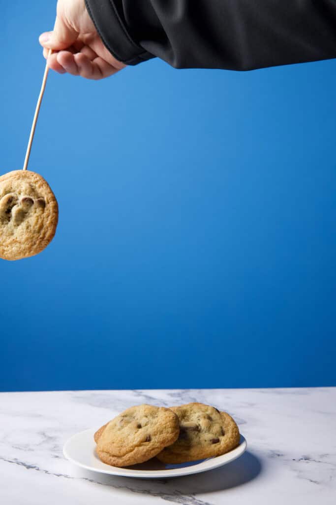 Single chocolate chip cookie photographed midair on a wooden skewer, held at an angle, positioned above a plate to be composited into a levitation image.