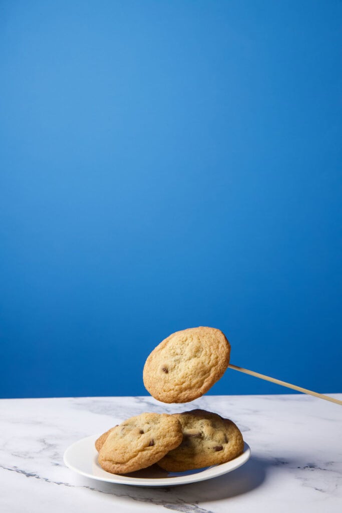 Single chocolate chip cookie photographed midair on a wooden skewer, held much closer to the plate to be composited into a levitation image.